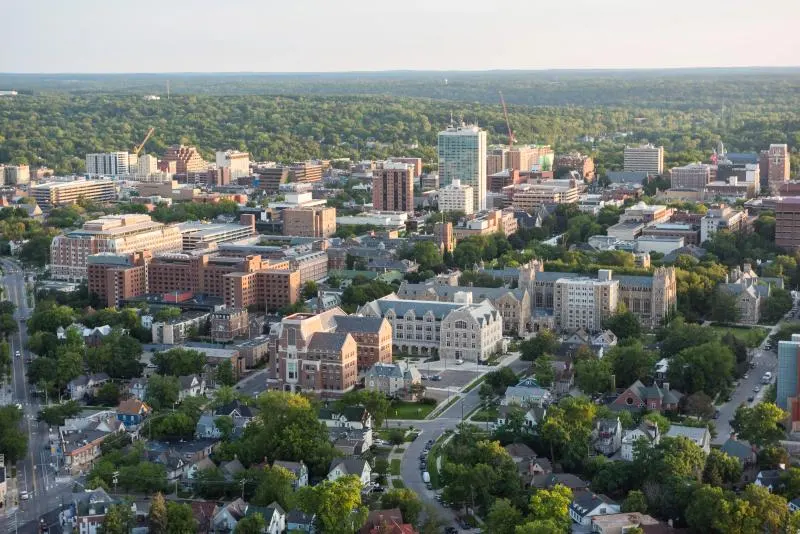 An aerial view of the University of Michigan's Ann Arbor campus at golden hour, showing a mix of Gothic and modern university buildings nestled among mature trees, with the surrounding city neighborhood visible in the foreground.